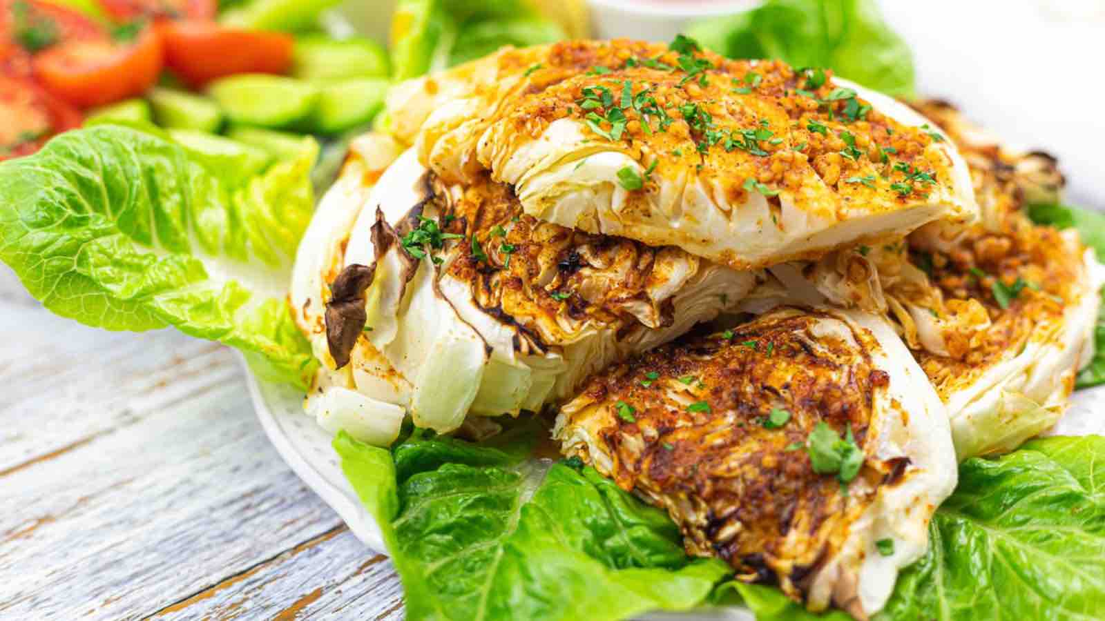 Grilled cabbage wedges topped with herbs served on a bed of lettuce, with sliced cucumbers and tomatoes in the background.