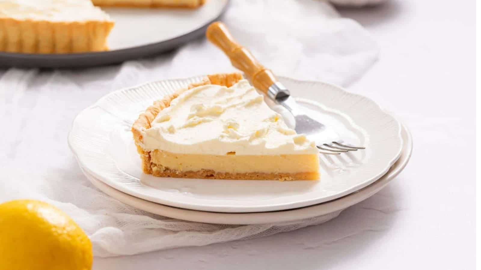A slice of lemon cream pie topped with whipped cream sits on a white plate with a dessert fork; a lemon is visible in the foreground.