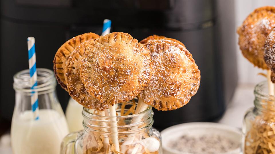 Round, golden-brown pie pops on sticks are displayed in mason jars, dusted with powdered sugar, with bottles of milk and striped straws nearby.
