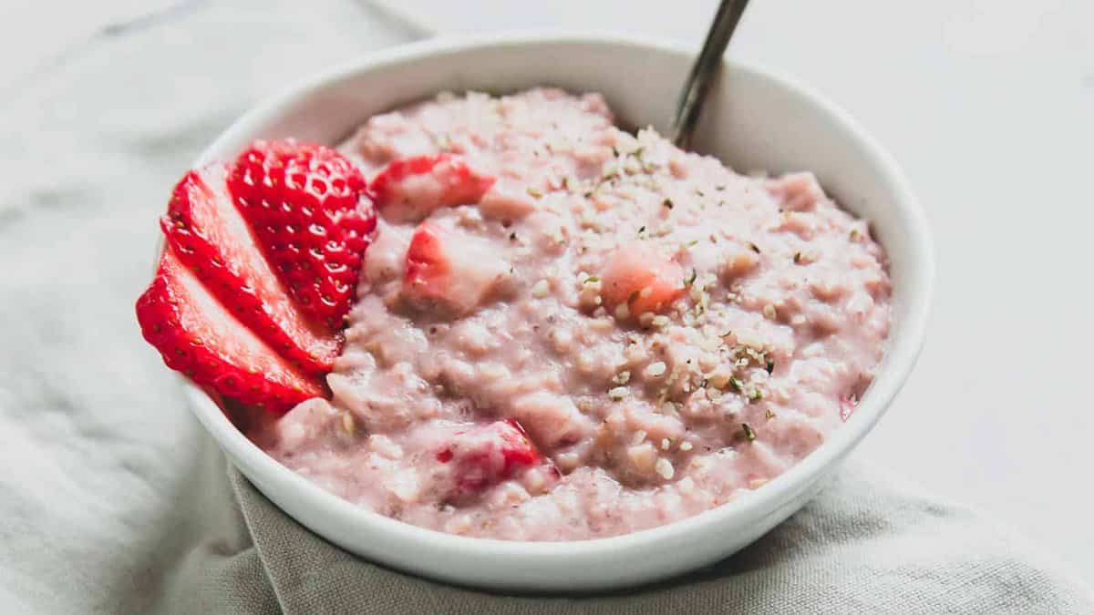 A bowl of strawberry oatmeal, topped with sliced strawberries and sprinkled seeds, with a spoon in the bowl.