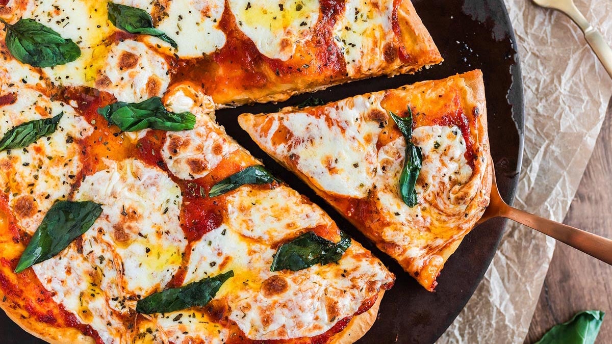A Margherita pizza with melted mozzarella, tomato sauce, and basil leaves, with one slice being lifted from the whole pizza on a dark round tray.