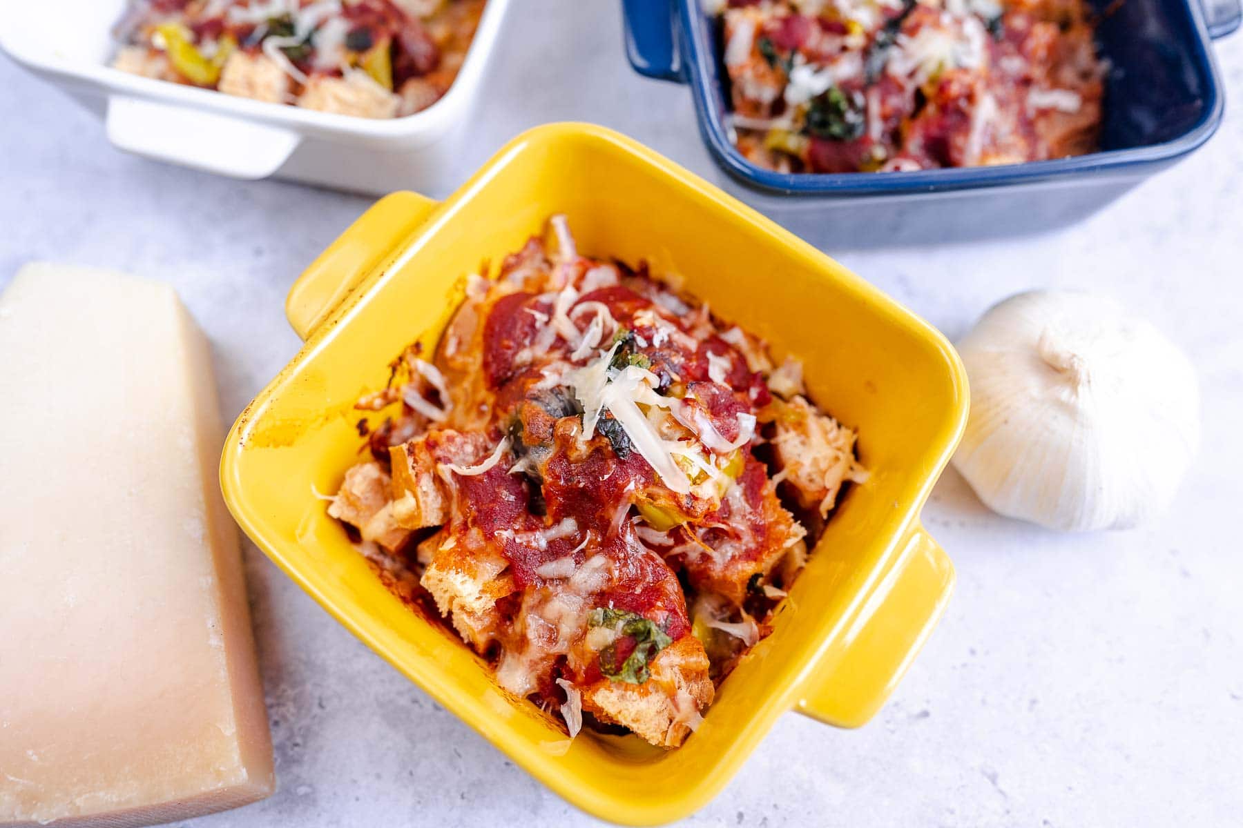 A yellow dish filled with baked casserole topped with tomato sauce and shredded cheese, next to a garlic bulb and a block of cheese.