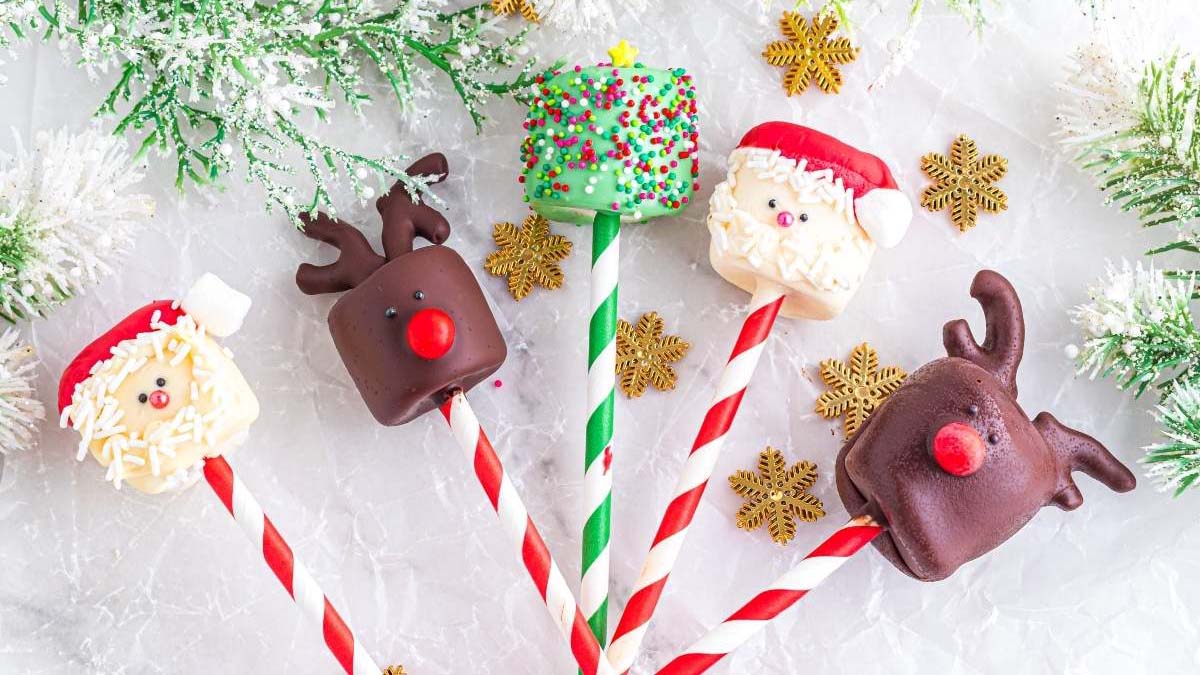 Five decorated Christmas marshmallow pops on striped sticks are arranged on a white surface with fake snow, pine branches, and gold snowflake ornaments.