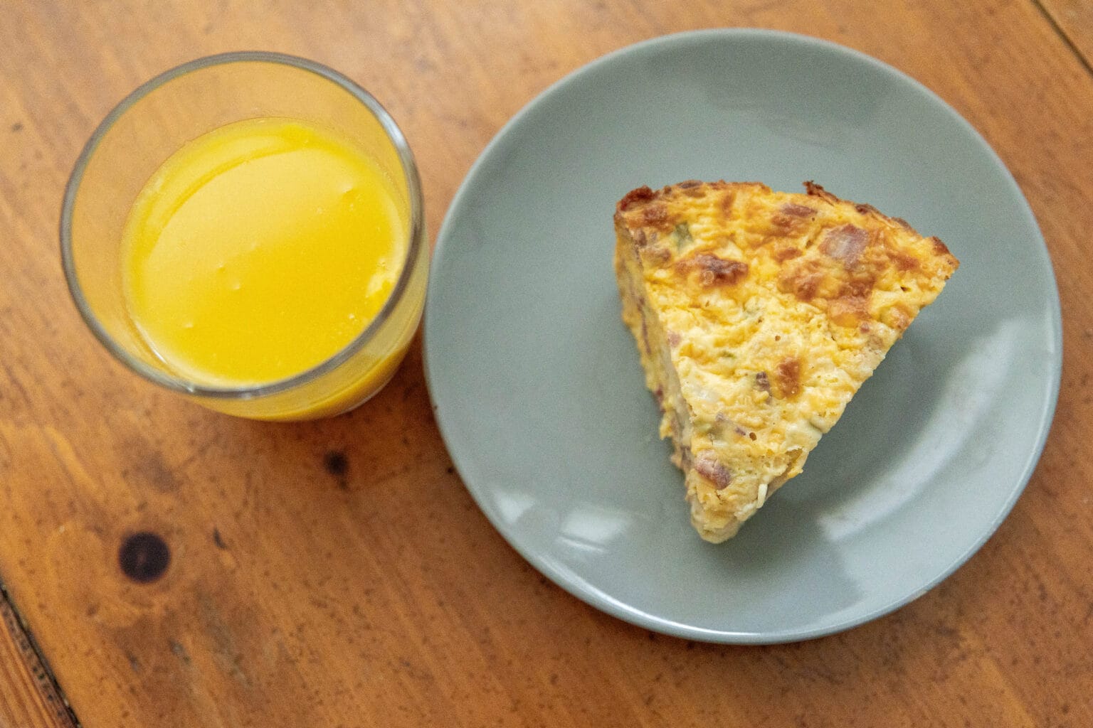 A glass of orange juice and a slice of egg casserole with ham on a gray plate, set on a wooden table.