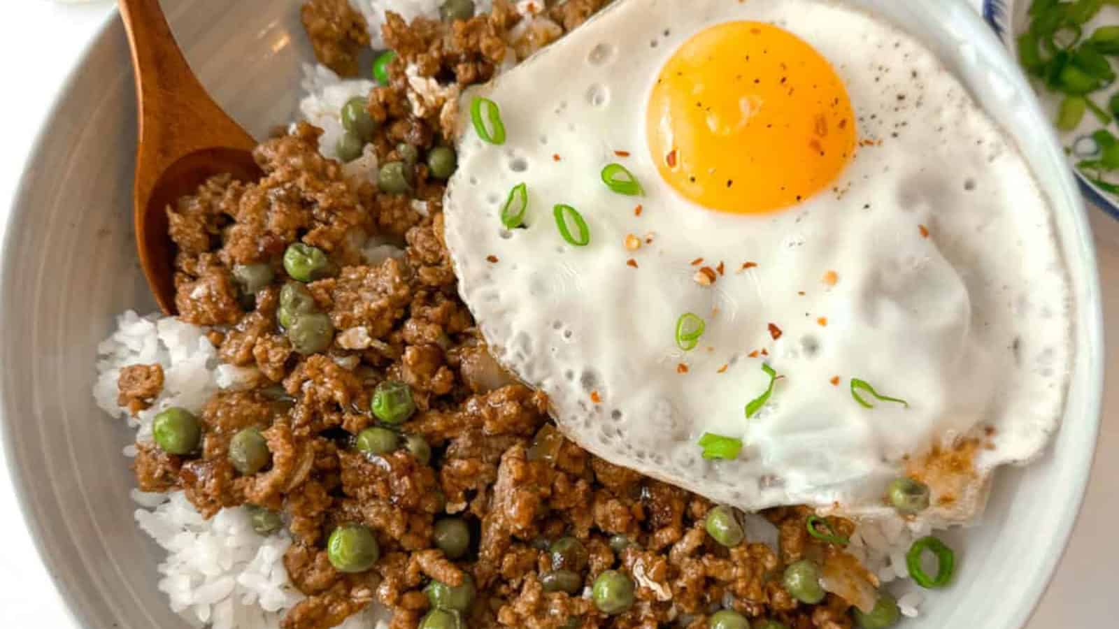 A bowl of white rice topped with ground meat, green peas, and a sunny-side-up egg, garnished with chopped green onions.