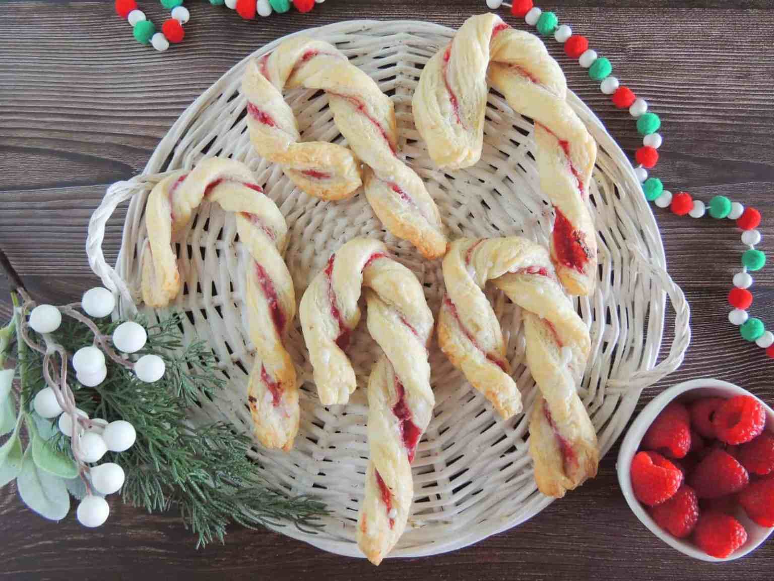 Six puff pastry sticks shaped like candy canes with red filling on a white woven plate, decorated with greenery, white berries, and a bowl of strawberries.
