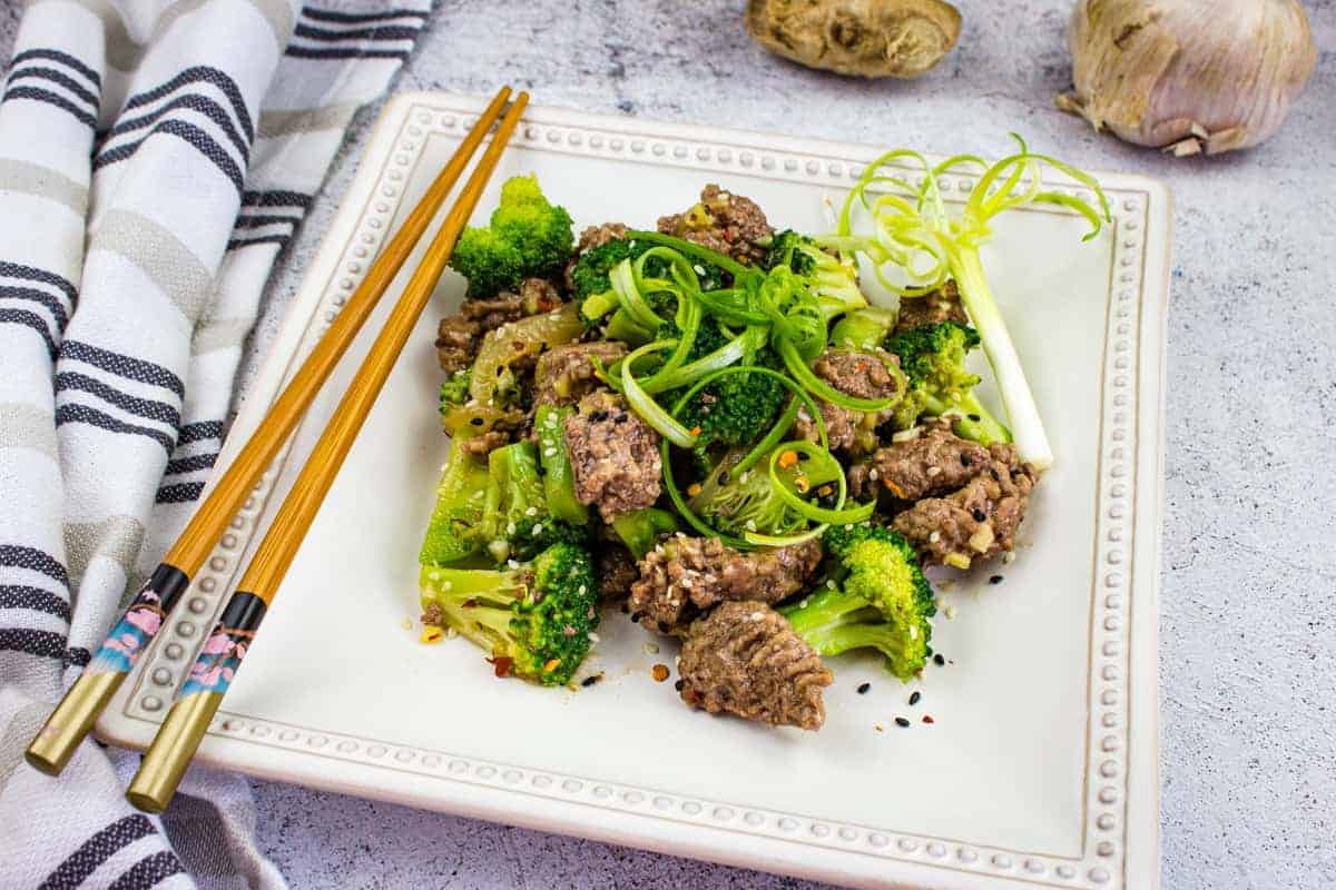 A square plate with beef and broccoli stir-fry garnished with sliced green onions, next to a pair of chopsticks on a striped napkin. Ginger and garlic bulbs are in the background.
