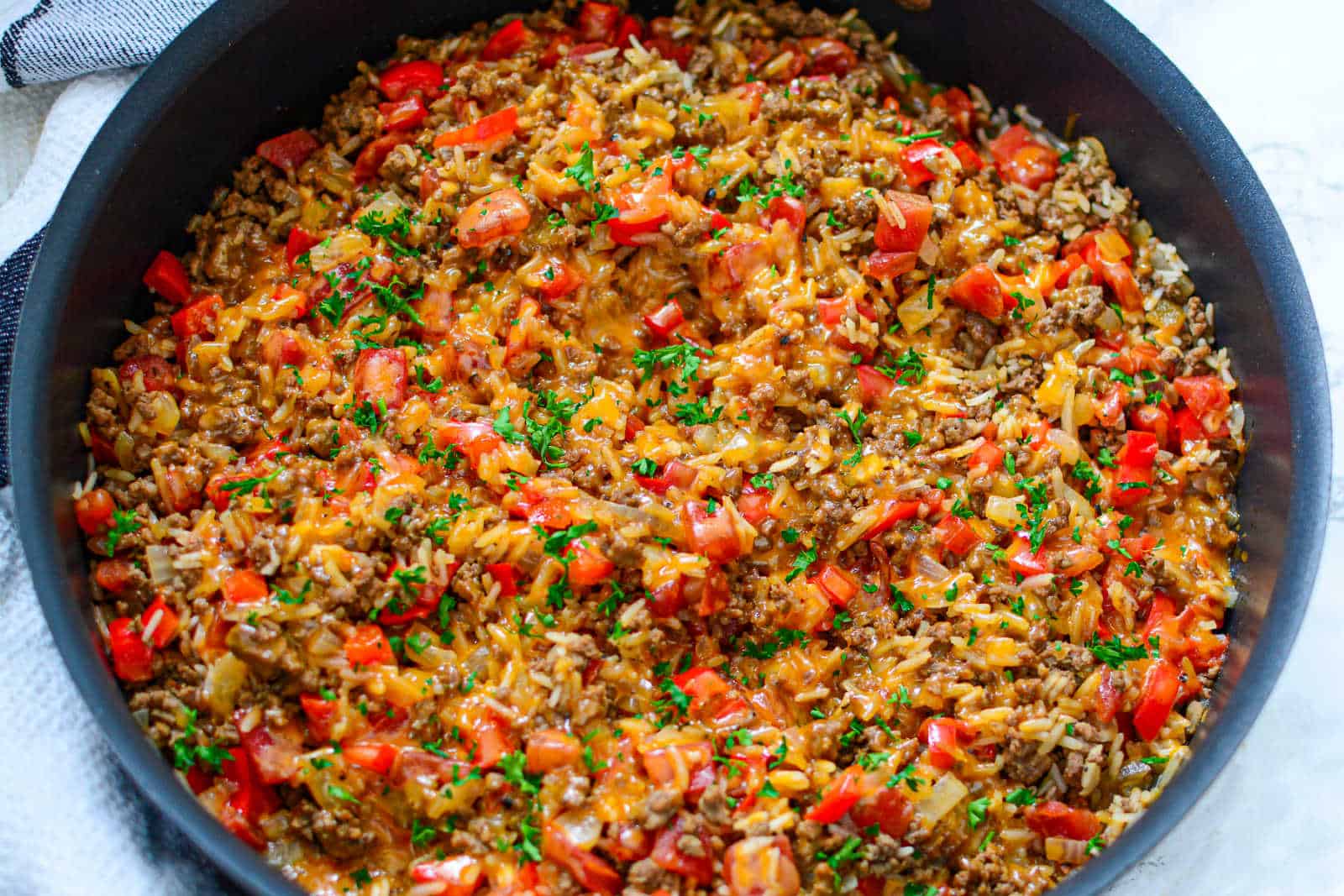 A skillet filled with a mixture of cooked ground beef, rice, melted cheese, red bell peppers, and chopped parsley.