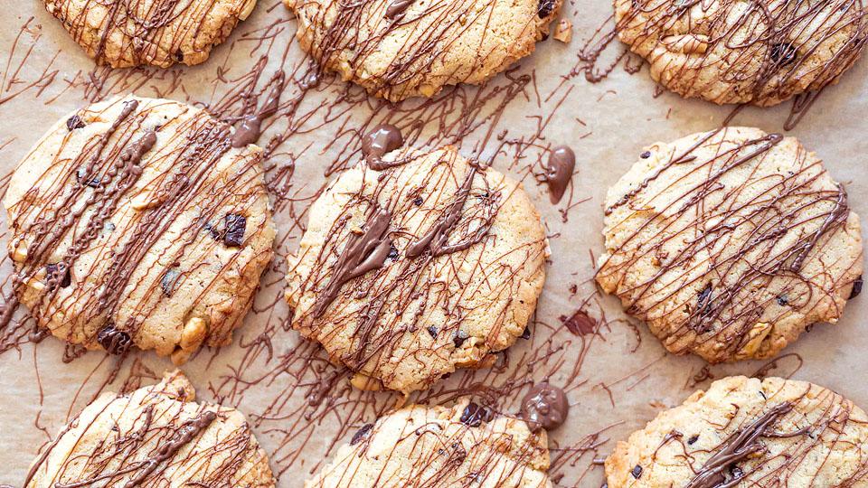 Nine cookies with chocolate drizzle on parchment paper. The cookies contain visible pieces of nuts and possibly chocolate chips or raisins.