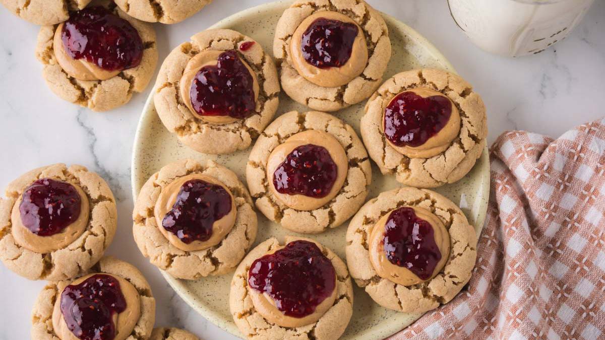 A plate of peanut butter cookies topped with dollops of red jelly, with additional cookies on the side and a checkered cloth partially visible.
