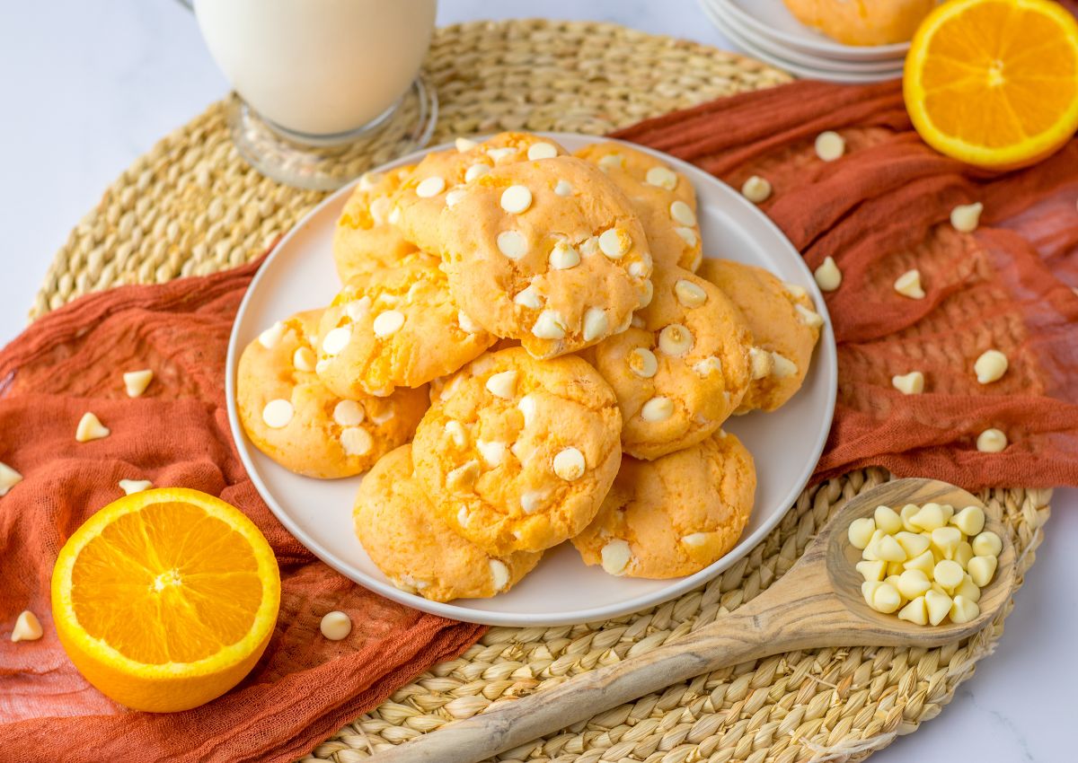 A plate of orange cookies with white chocolate chips, surrounded by orange slices and a wooden spoon with chocolate chips, on a woven mat with a glass of milk.