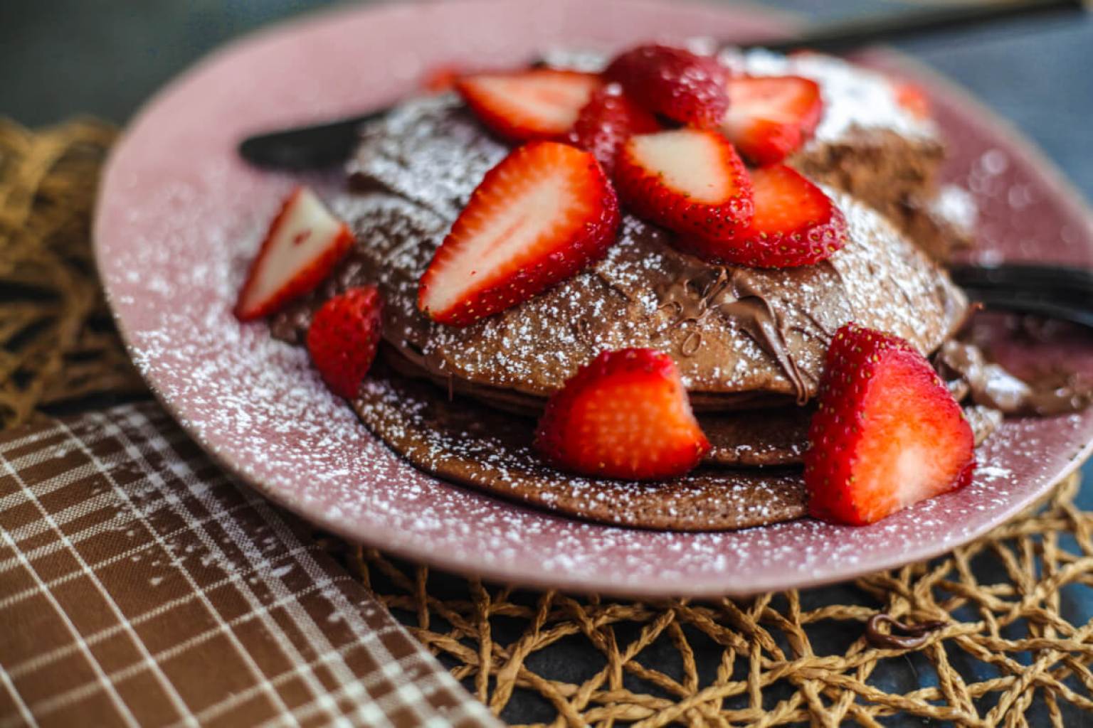 A plate of chocolate pancakes topped with sliced strawberries and a dusting of powdered sugar, placed on a woven mat next to a plaid napkin.