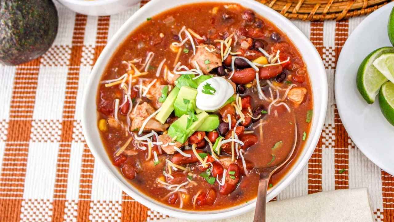 A bowl of chili topped with shredded cheese, avocado, sour cream, and cilantro, served with a spoon on a checkered tablecloth. Lime wedges are on the side.