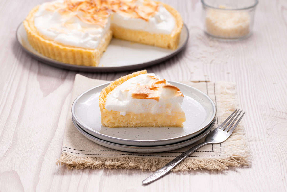 A slice of coconut cream pie with toasted coconut topping sits on a plate, with the remaining pie in the background. A fork is placed next to the plate on a napkin.