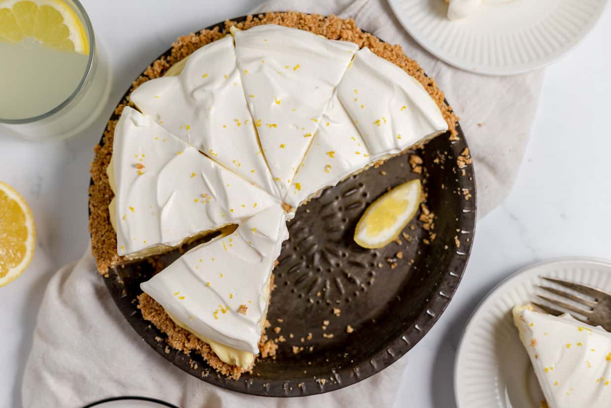 A lemon cream pie with a graham cracker crust, topped with whipped cream and lemon zest, sits on a table with several slices cut and served on plates.