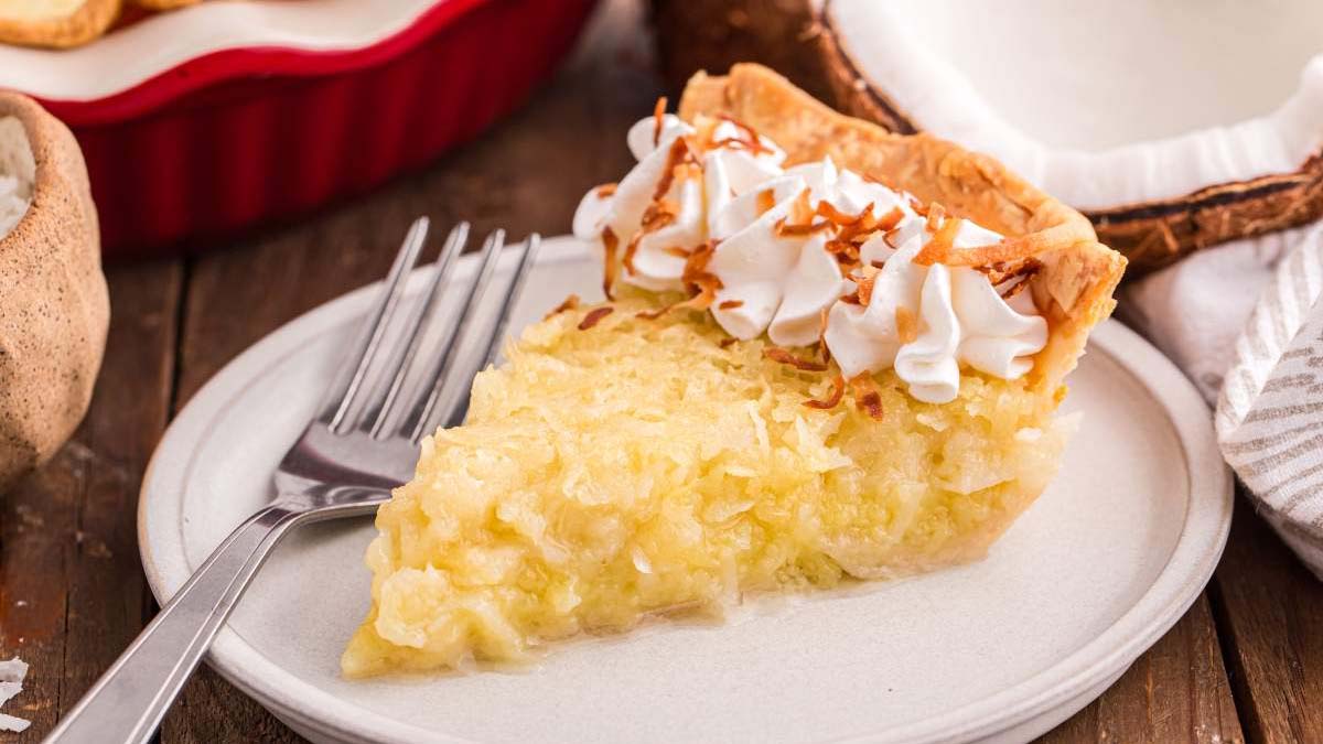 A slice of coconut pie topped with whipped cream and toasted coconut sits on a plate with a fork.