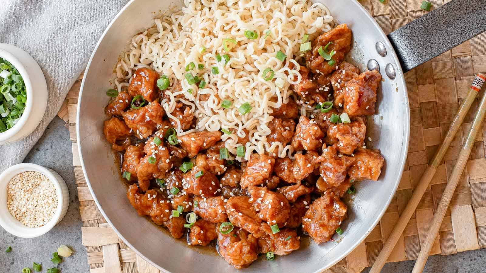 A skillet containing cooked ramen noodles and pieces of saucy chicken, garnished with sliced green onions and sesame seeds, sits on a woven placemat with chopsticks nearby.