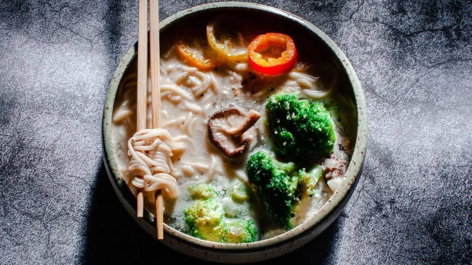 A bowl of noodle soup with broccoli, mushrooms, bell pepper slices, and chopsticks resting on the rim.
