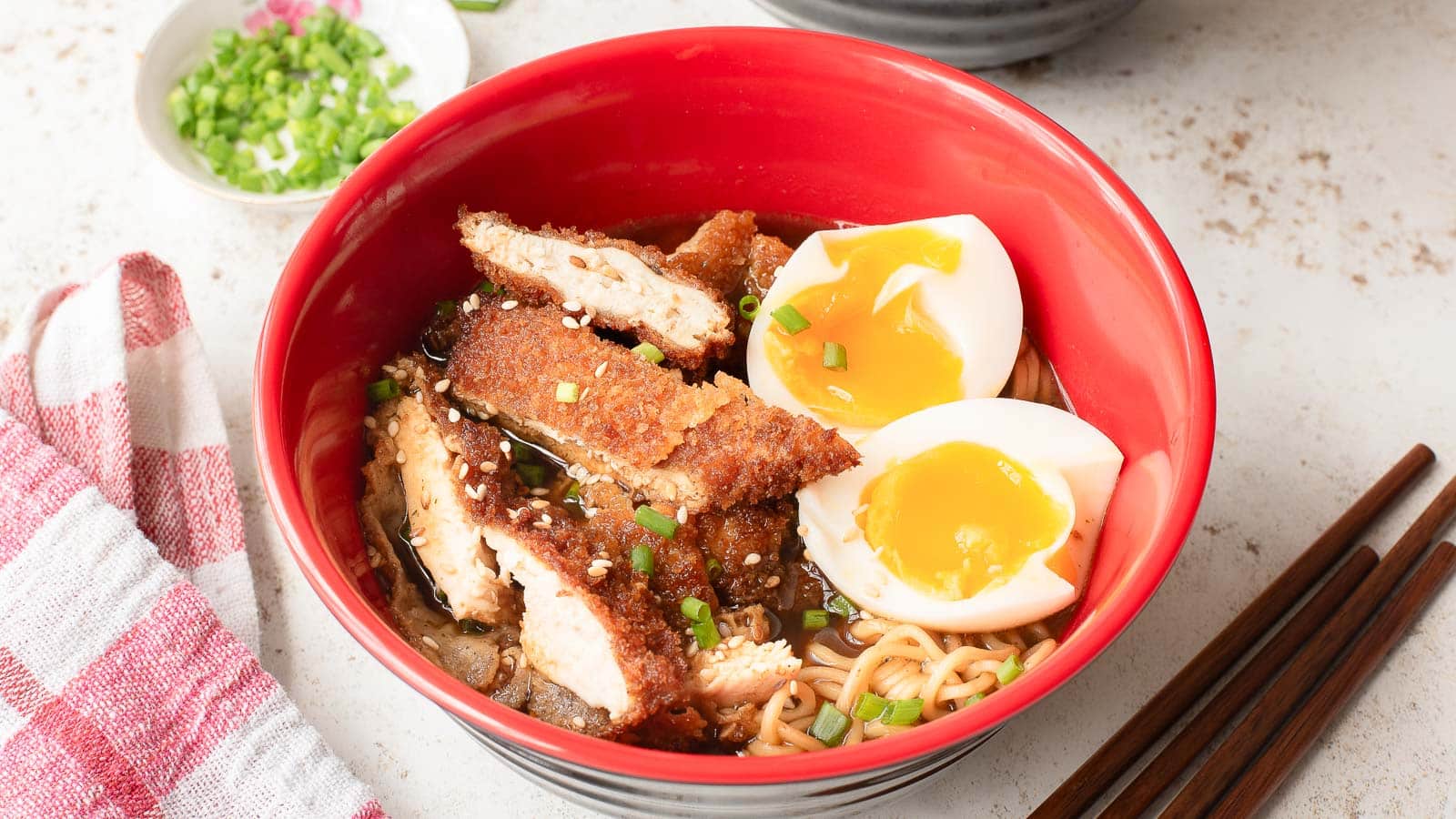 A red bowl filled with ramen noodles, sliced breaded chicken, two halved soft-boiled eggs, chopped green onions, and sesame seeds, with chopsticks beside it.