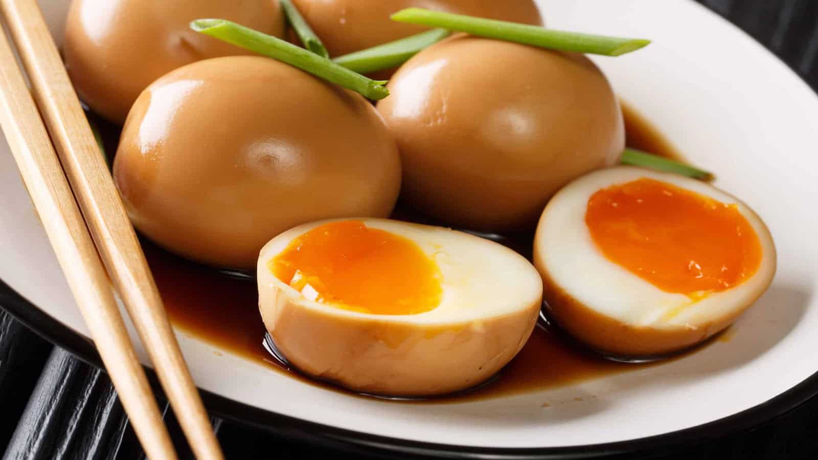 A plate of marinated soft-boiled eggs with runny yolks, garnished with green onions and served with soy sauce, and a pair of chopsticks beside the plate.