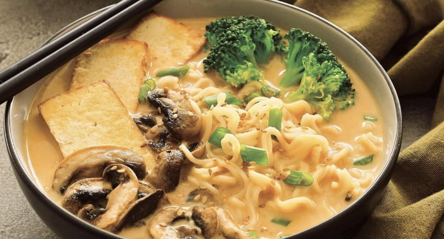 A bowl of ramen noodles in broth, topped with sliced tofu, mushrooms, broccoli florets, and chopped green onions, with a pair of black chopsticks resting on the bowl.