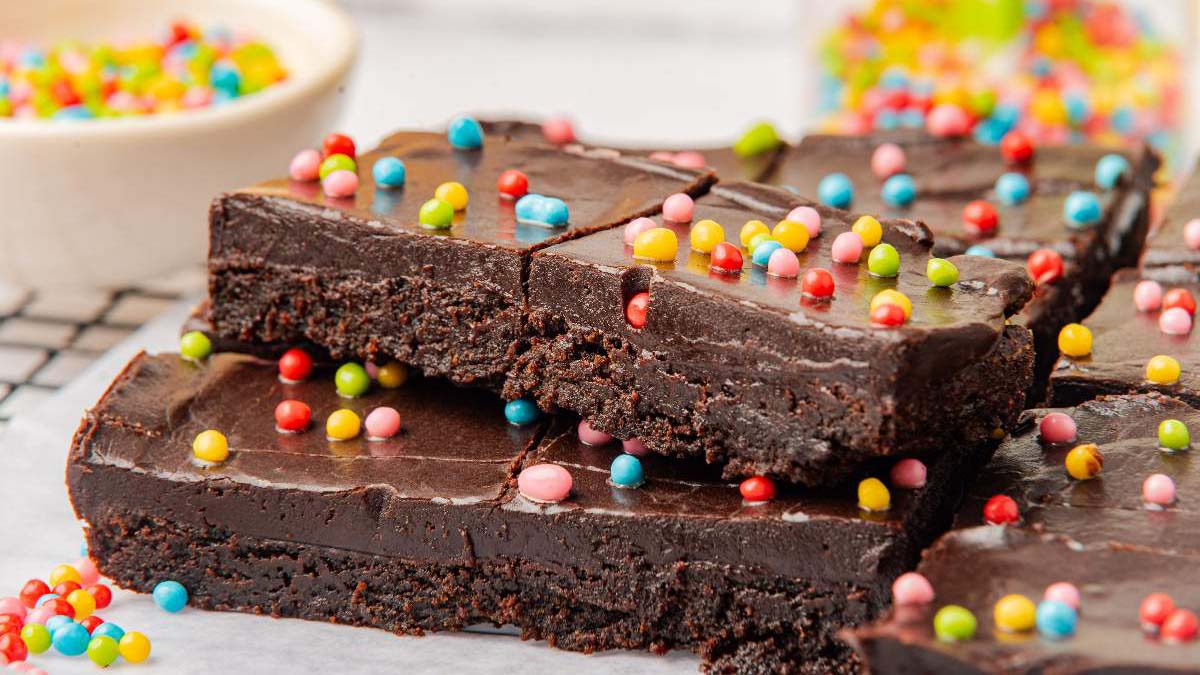 Three chocolate brownies are stacked on a surface and topped with colorful round candy sprinkles. More sprinkles are scattered around.