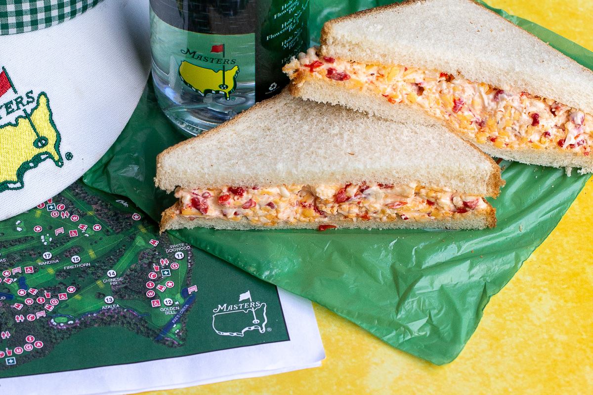 A pimento cheese sandwich on white bread is cut in half, placed on a green wrapper next to a Masters-branded water bottle, a hat, and a map of the golf course.