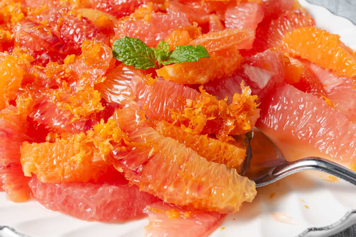 Close-up of grapefruit and orange segments with orange zest and a mint garnish on a white plate, with a serving spoon.