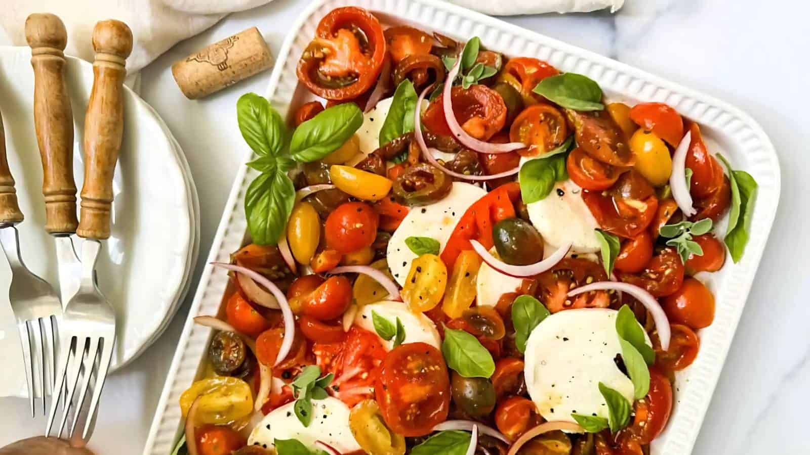 A rectangular plate of Caprese salad with sliced tomatoes, mozzarella, fresh basil, red onions, and a sprinkle of black pepper. Plates, forks, and a cork are beside the dish.