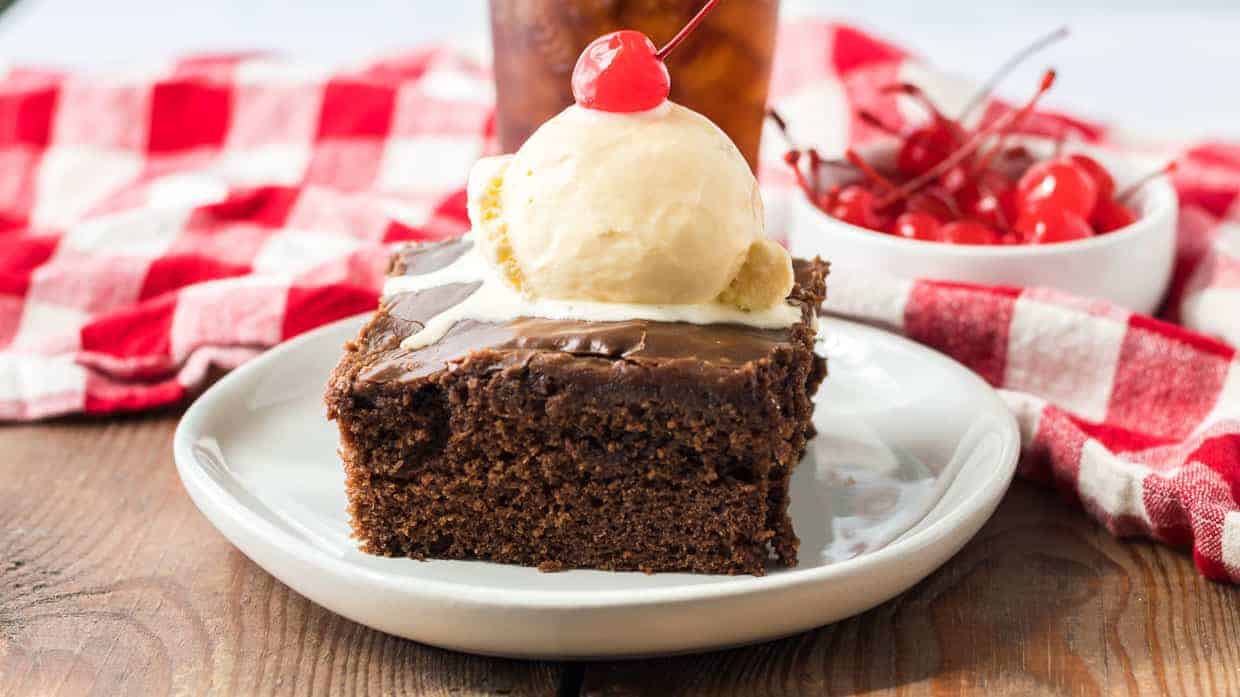 A square piece of chocolate cake topped with a scoop of vanilla ice cream and a cherry, served on a white plate with a red and white checkered cloth in the background.