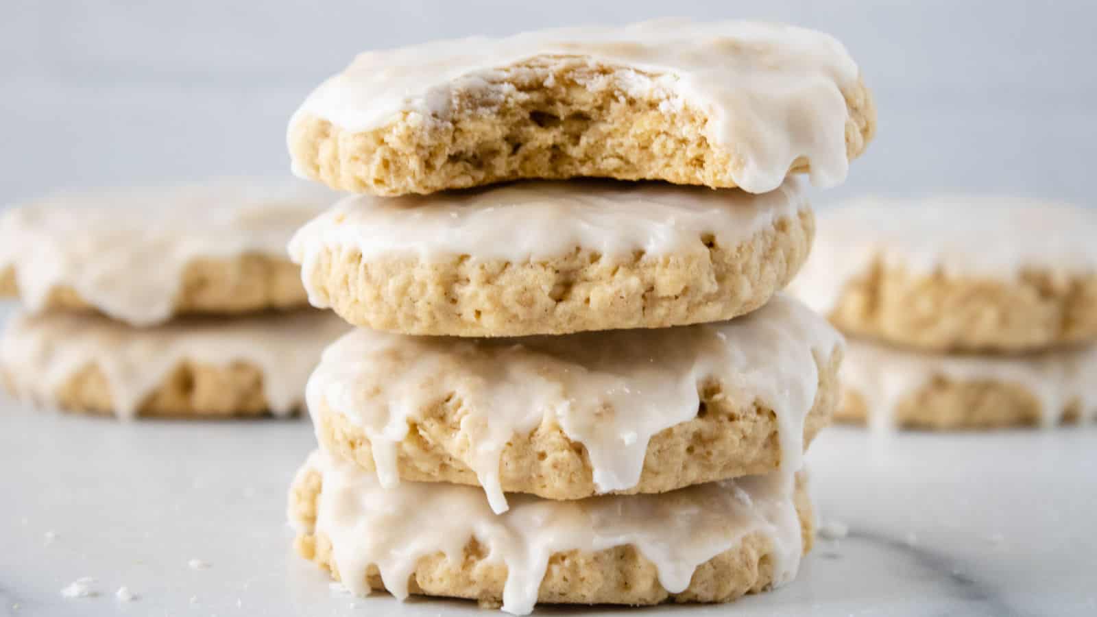 Three stacked iced cookies with a bite taken out of the top one, with more iced cookies blurred in the background on a white surface.