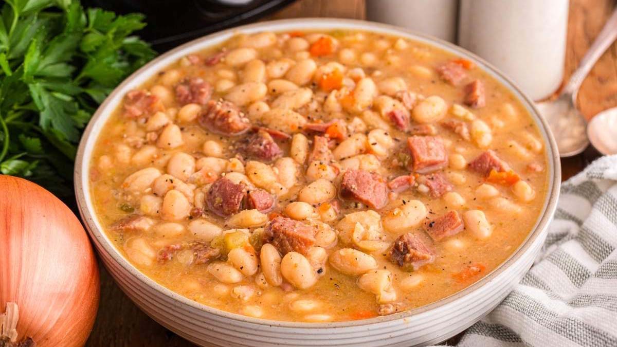 A bowl of white bean and ham soup with visible pieces of ham, beans, and vegetables, placed on a wooden table next to an onion and fresh parsley.