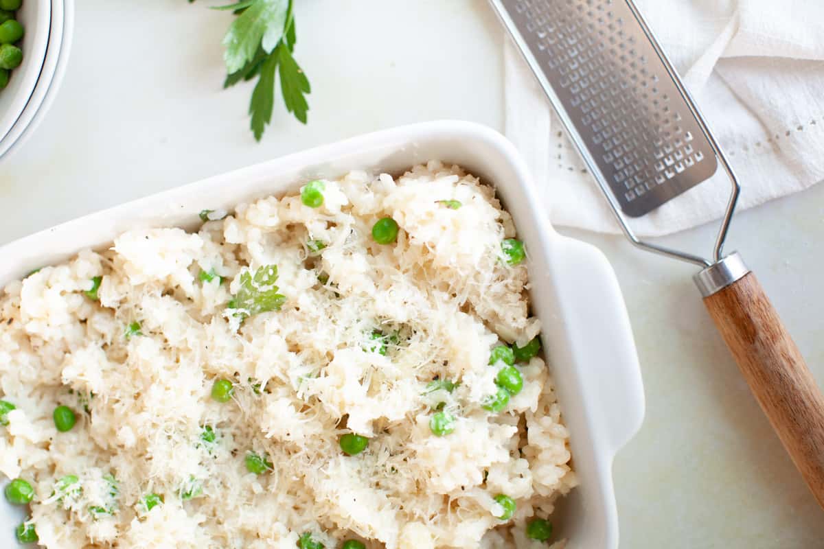 A white baking dish filled with risotto topped with grated cheese and green peas, next to a metal grater and parsley on a light surface.