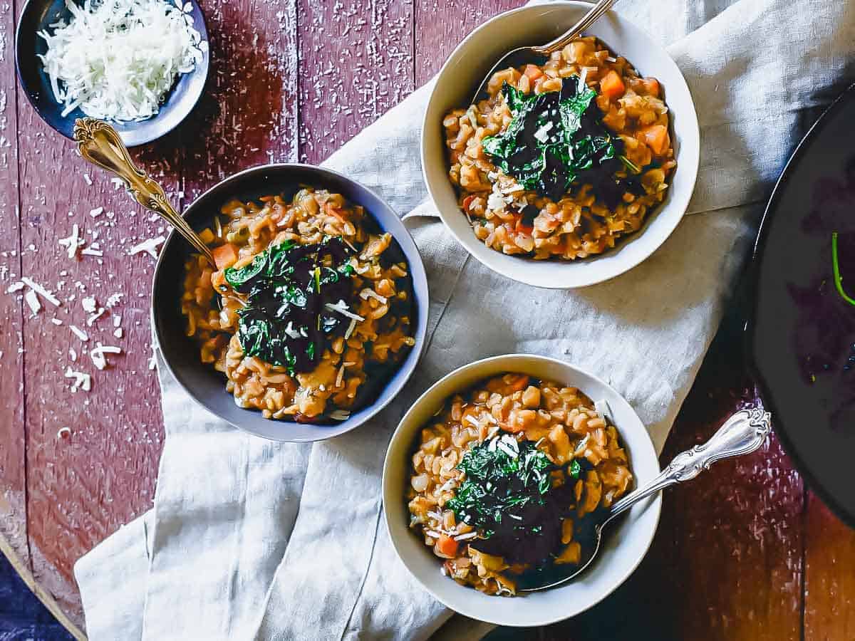 Three bowls of risotto topped with chopped herbs, placed on a cloth over a wooden table, with a small bowl of grated cheese nearby.