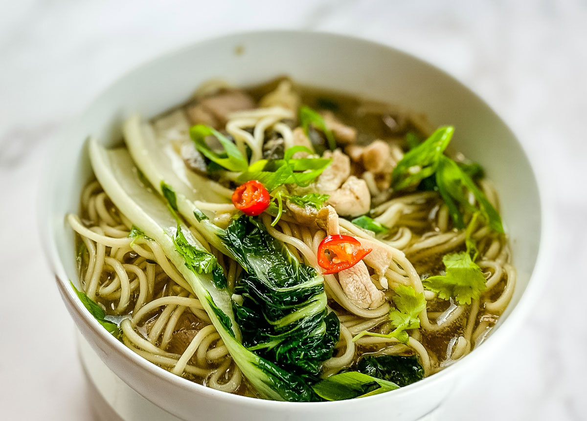 A bowl of noodle soup with bok choy, chicken slices, chopped green onions, and red chili peppers in a light broth.
