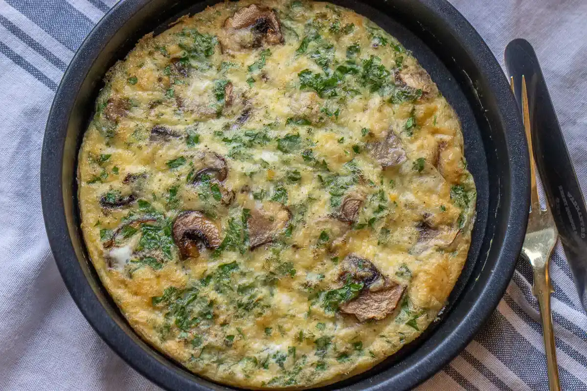 A baked frittata in a round pan, featuring visible pieces of mushrooms and chopped green herbs, placed on a striped cloth next to a fork and knife.