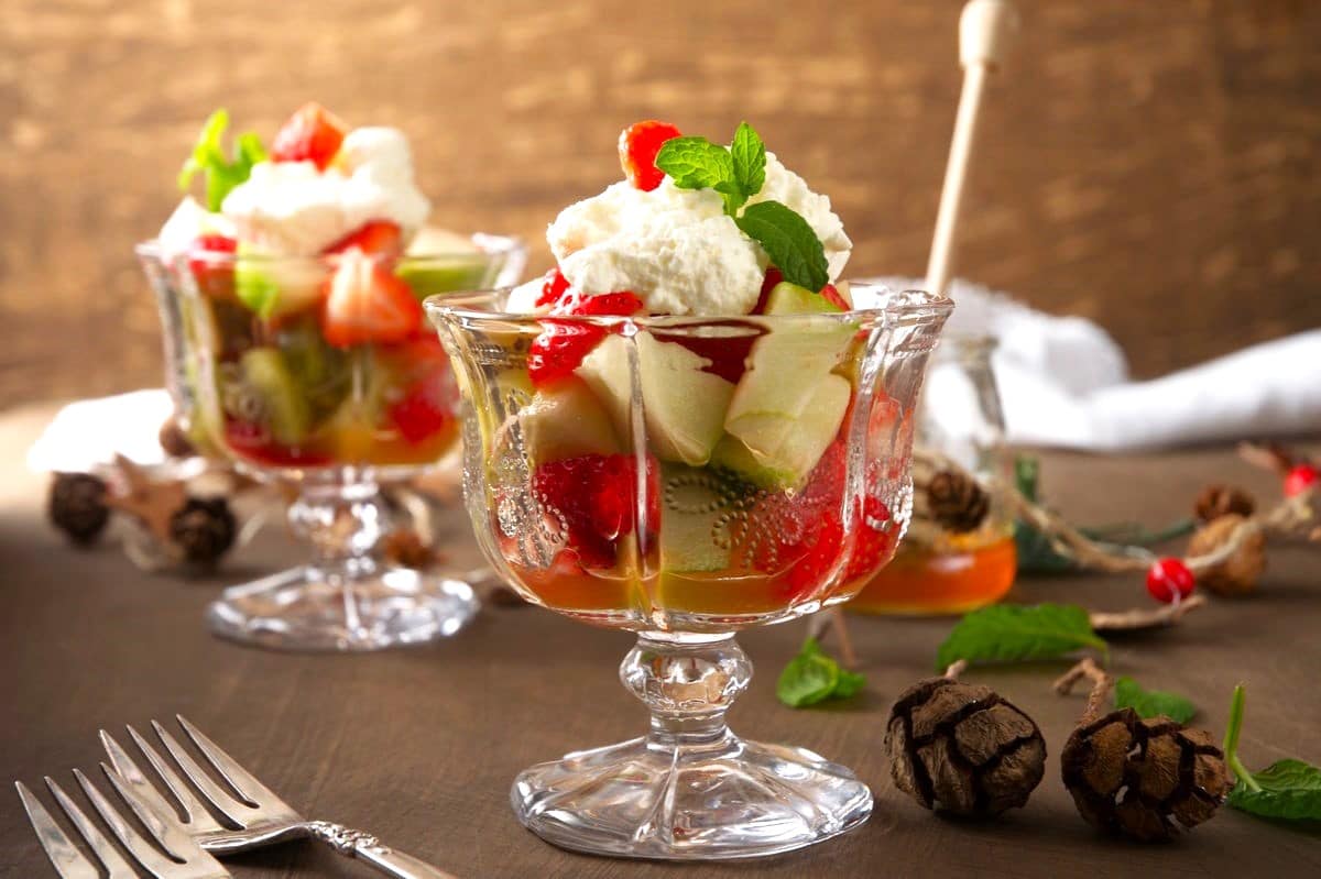 Two glass dessert cups filled with sliced kiwi, strawberries, syrup, and topped with whipped cream and mint leaves, placed on a wooden surface with pinecones and forks nearby.