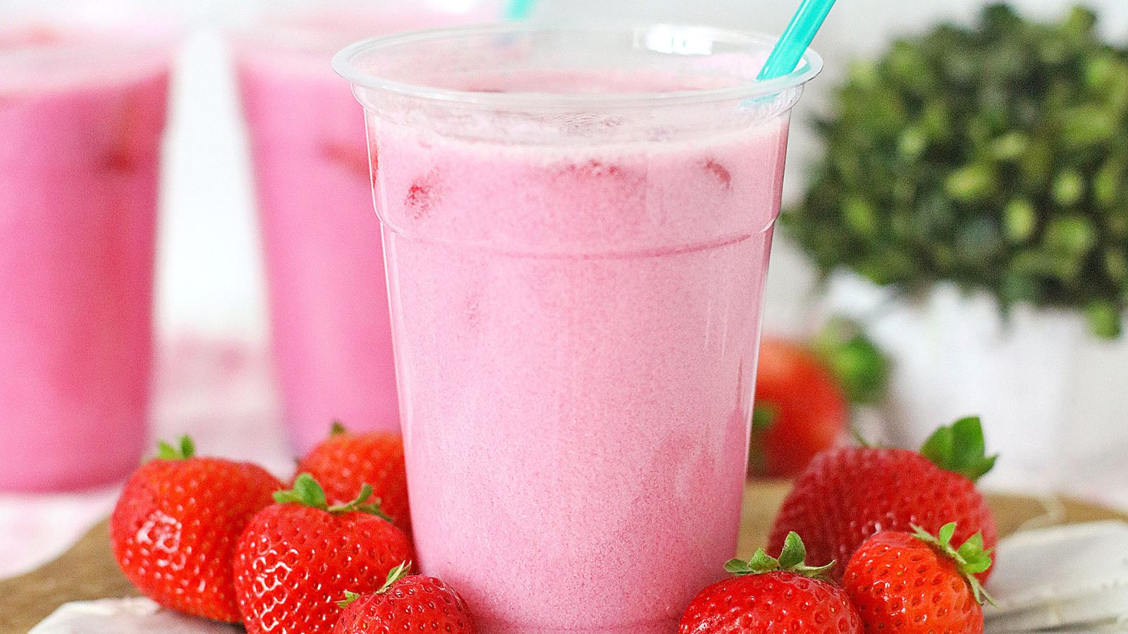 A clear plastic cup filled with a pink strawberry smoothie, surrounded by fresh strawberries, with a green straw in the cup.