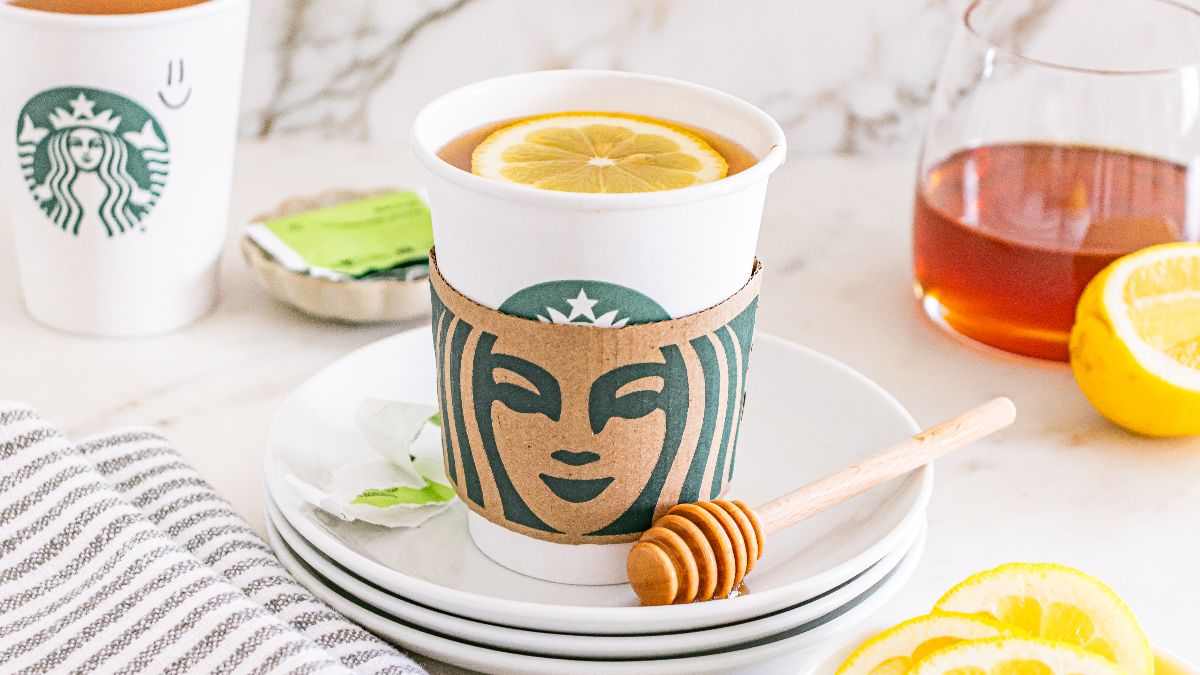 A cup of tea with a lemon slice on top, served in a Starbucks cup with a honey dipper, lemon slices, and honey pot nearby.
