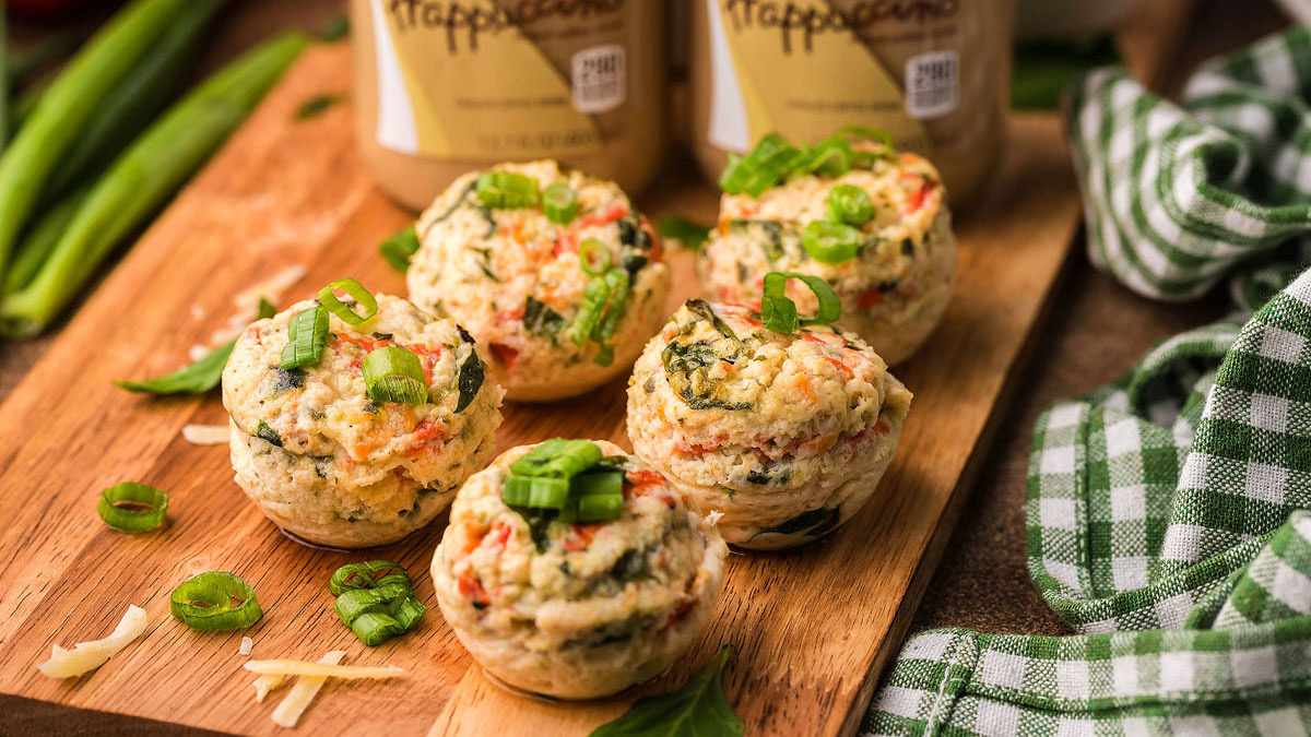 Five savory egg muffins with vegetables and green onions are arranged on a wooden board, with shredded cheese and green onions scattered nearby.