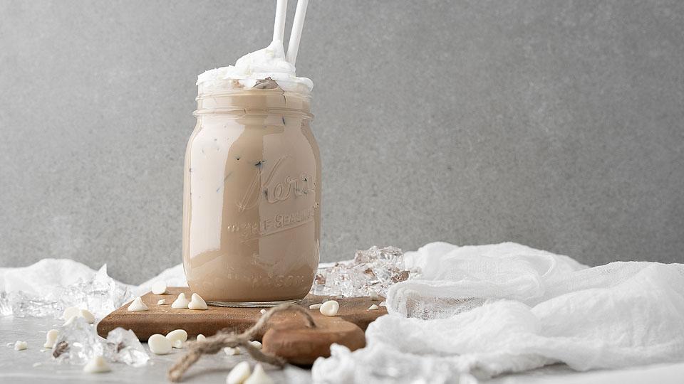 A mason jar filled with iced coffee and topped with whipped cream, two straws, ice cubes, and white chocolate chips on a wooden board with a white cloth nearby.