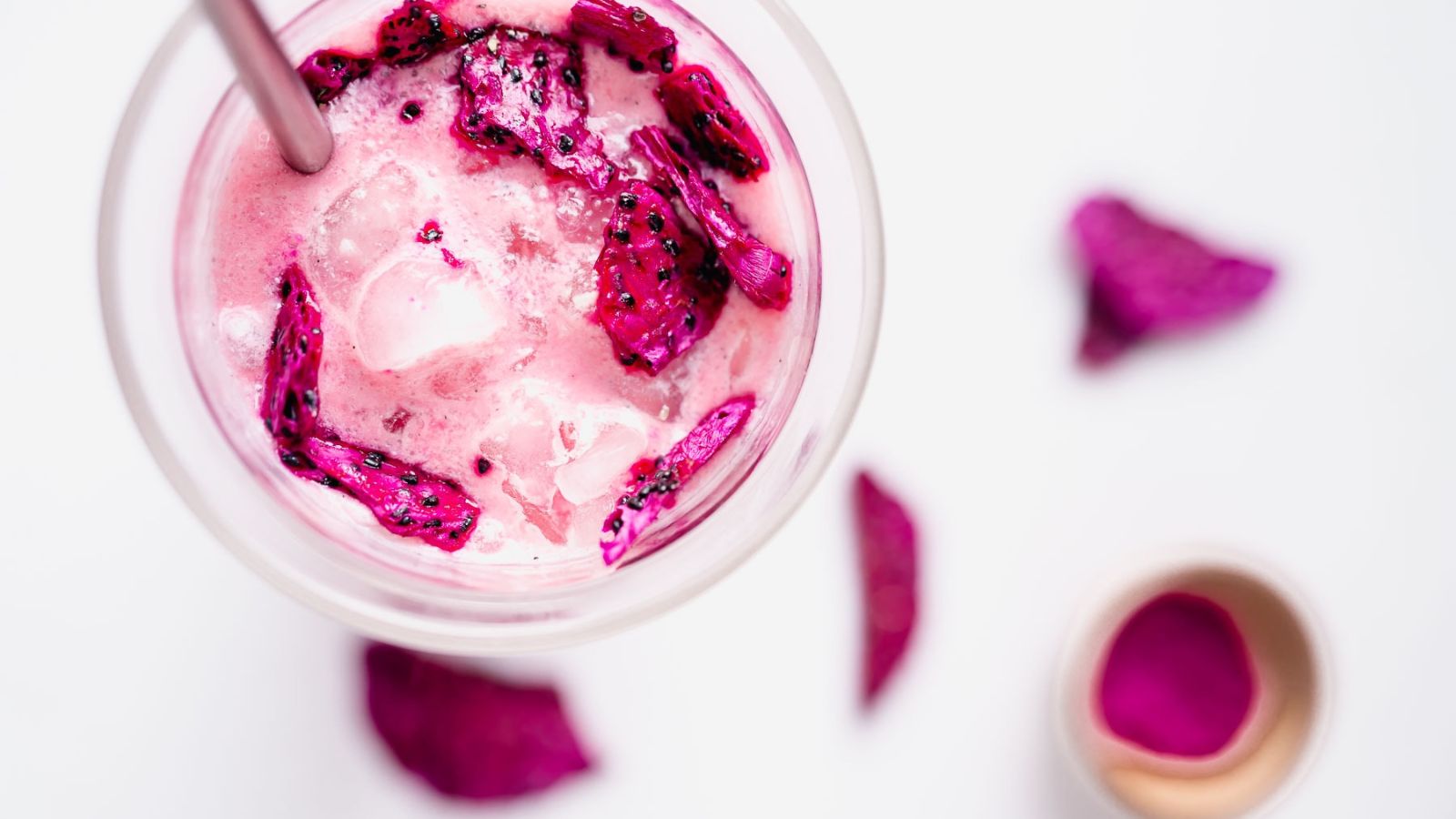 A glass of iced pink smoothie topped with dragon fruit pieces, with a metal straw, seen from above. Dragon fruit slices and a small cup are on the white surface nearby.