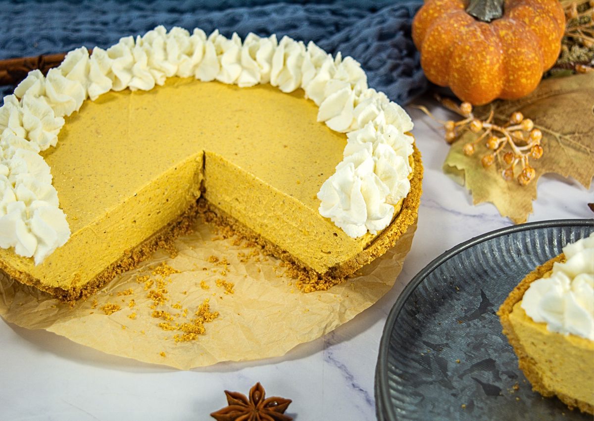 A pumpkin pie with a graham cracker crust and whipped cream topping, missing one slice, sits on parchment paper with fall decorations nearby.