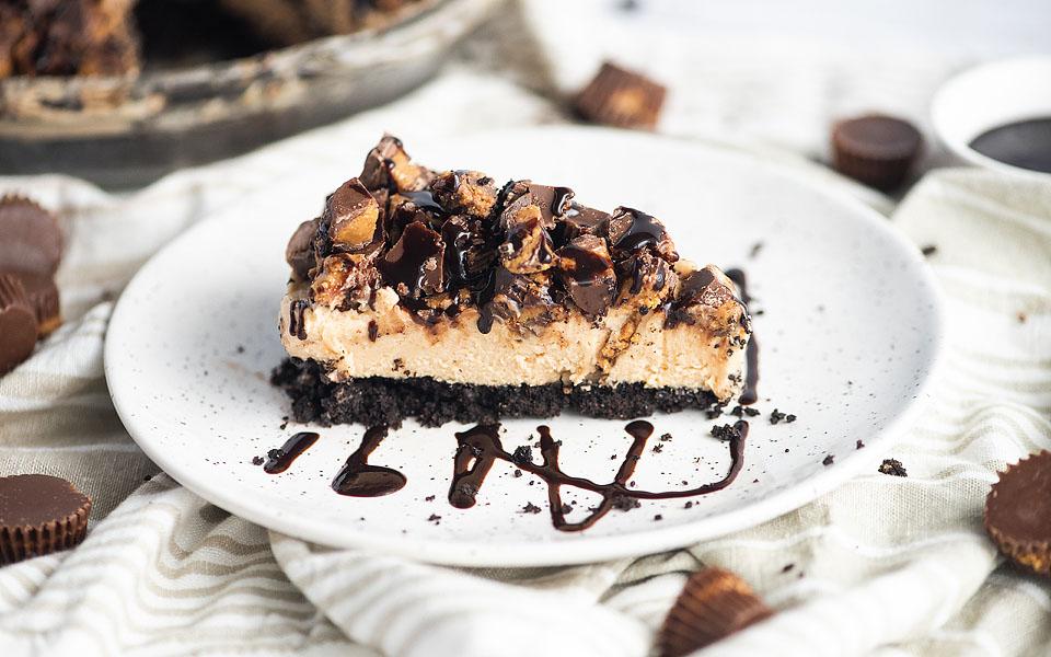 A slice of chocolate peanut butter pie with a cookie crust, topped with chopped peanut butter cups and drizzled with chocolate sauce on a white plate.