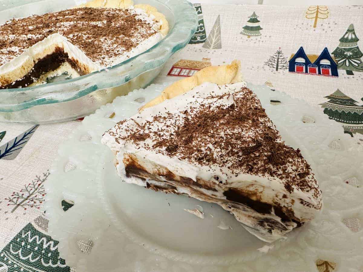 A slice of chocolate cream pie with whipped topping and chocolate shavings on a plate, with the remaining pie in a glass dish nearby on a festive tablecloth.