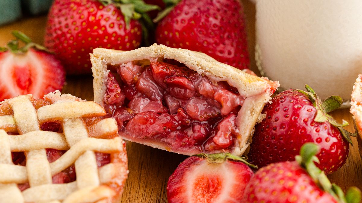 A close-up of a sliced mini strawberry pie with a lattice crust, fresh strawberries, and a glass of milk in the background.