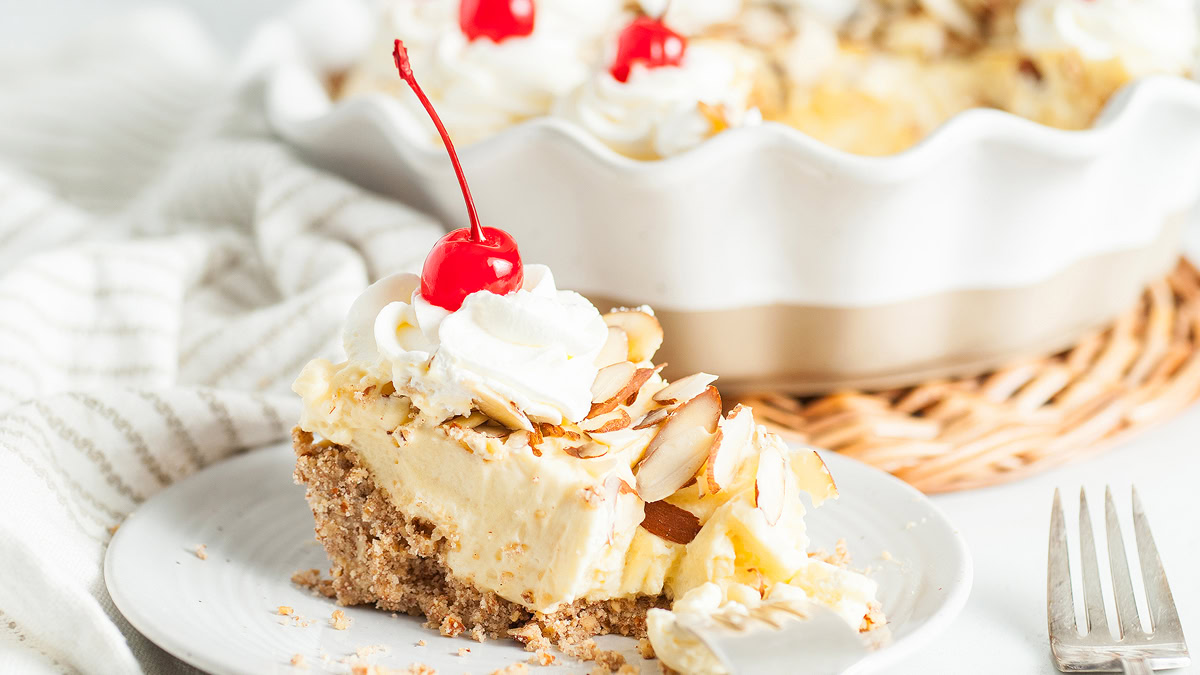 A slice of creamy pie topped with whipped cream, sliced almonds, and a cherry sits on a plate beside a fork, with the remaining pie in the background.