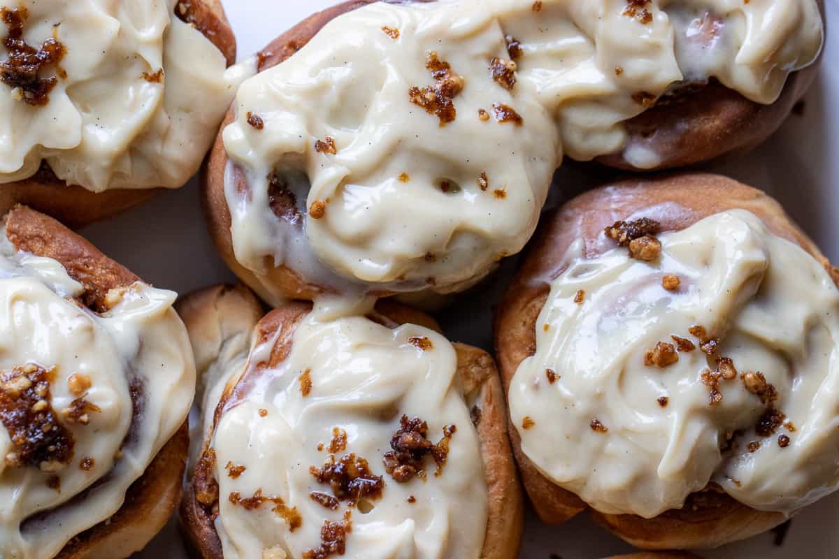 Close-up of several cinnamon rolls topped with thick cream cheese icing and sprinkled with small pieces of brown sugar or cinnamon.