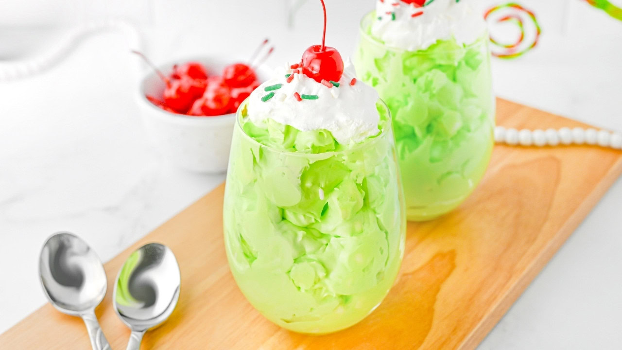 Two glasses of green pudding topped with whipped cream, sprinkles, and a cherry, served on a wooden tray with two spoons beside them. A bowl of cherries is in the background.