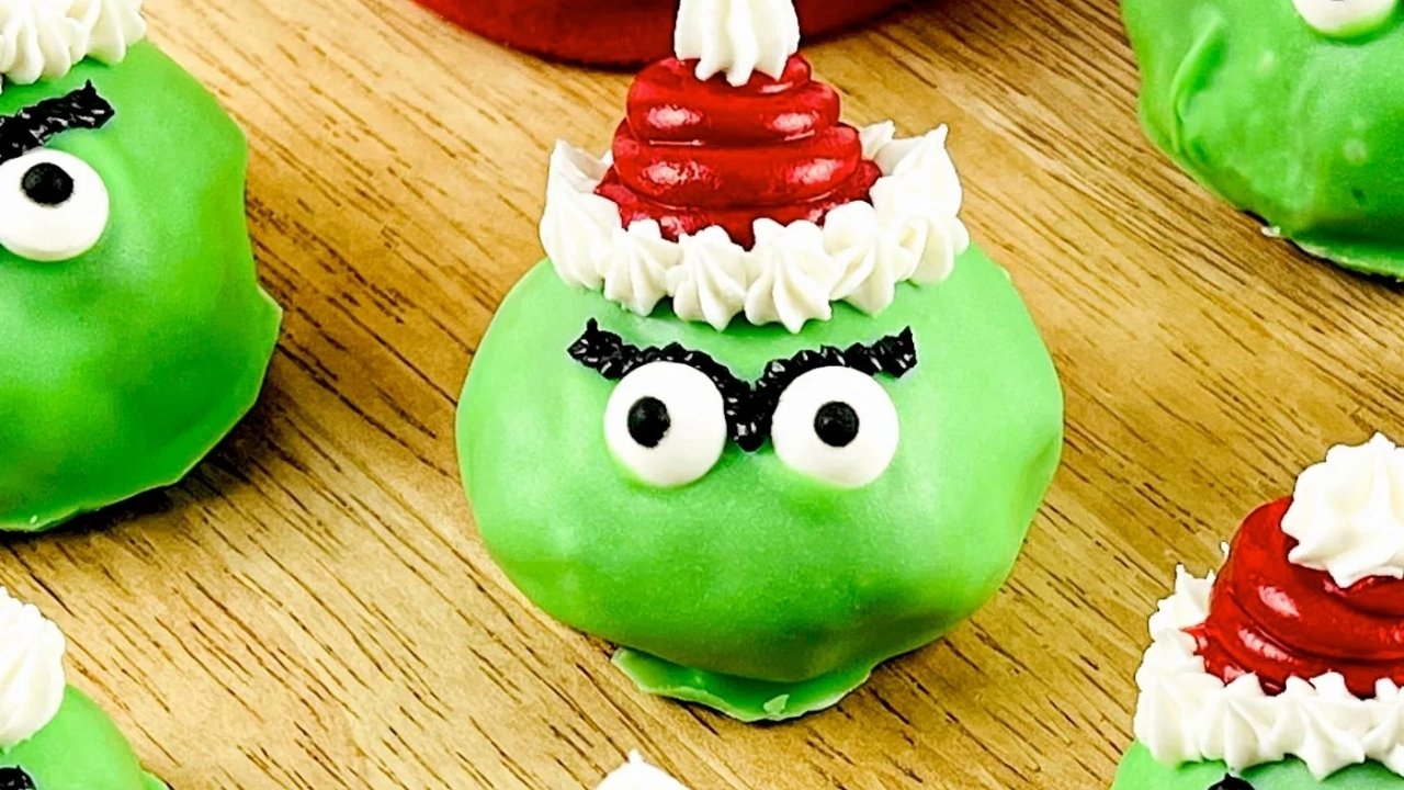 A green, round dessert decorated with candy eyes, black eyebrows, and a red and white icing Santa hat, placed on a wooden surface.